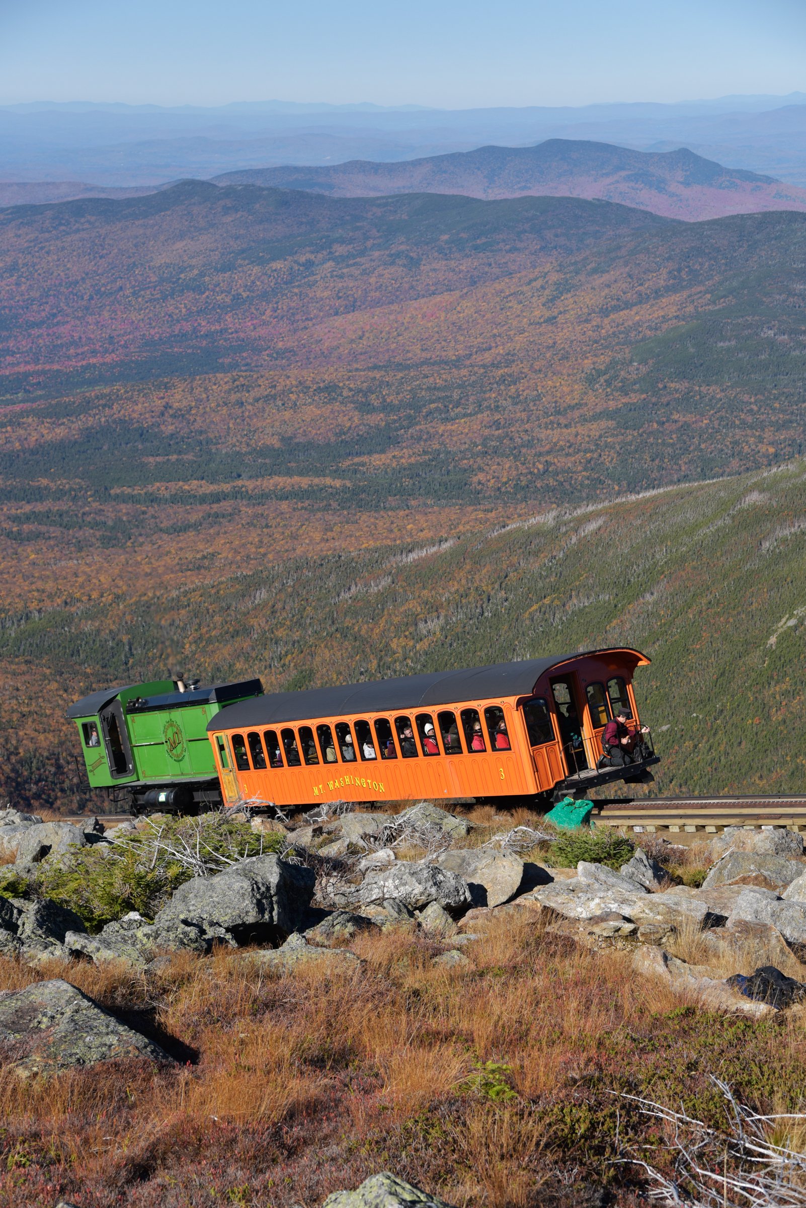 Orange and green Mount Washington Cog Railway train climbing through autumn mountain scenery
