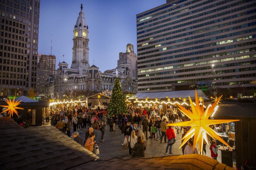 Philadelphia Christmas Village filled with visitors and illuminated stalls with City Hall behind it
