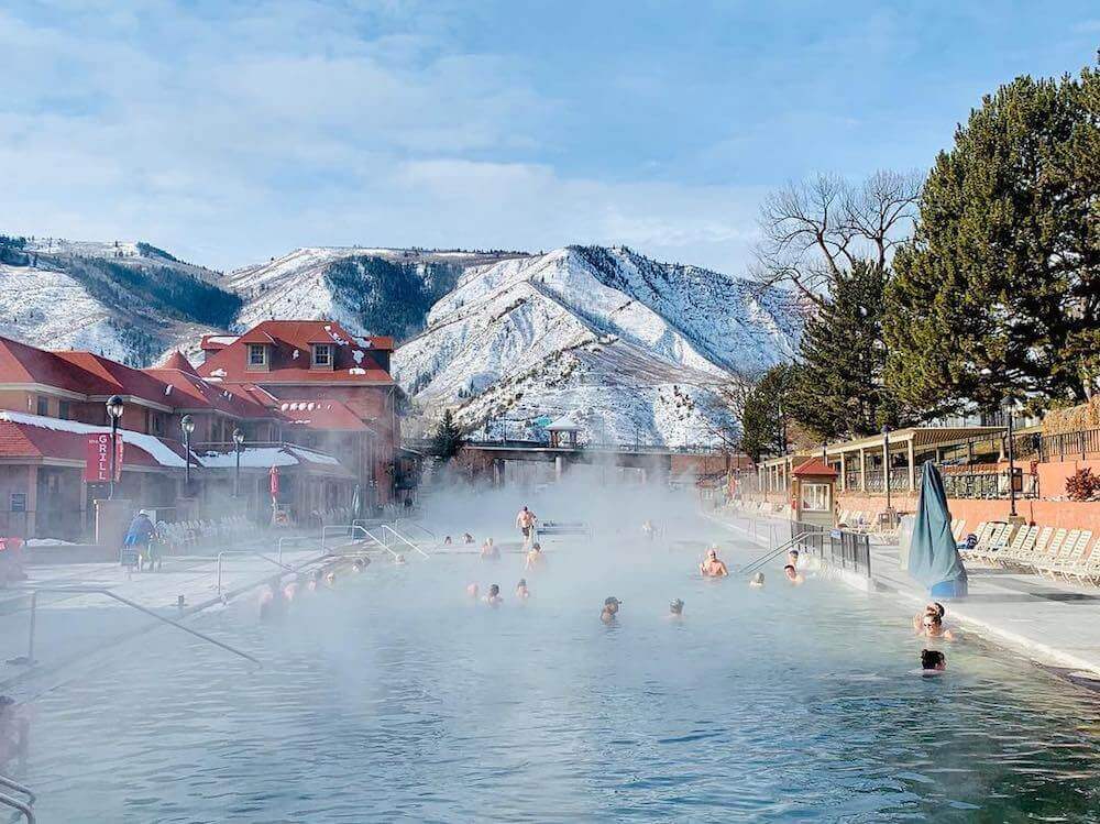 Guests soaking in a steaming outdoor pool at Glenwood Hot Springs Resort during winter, with snow-covered mountains behind the historic bathhouse.