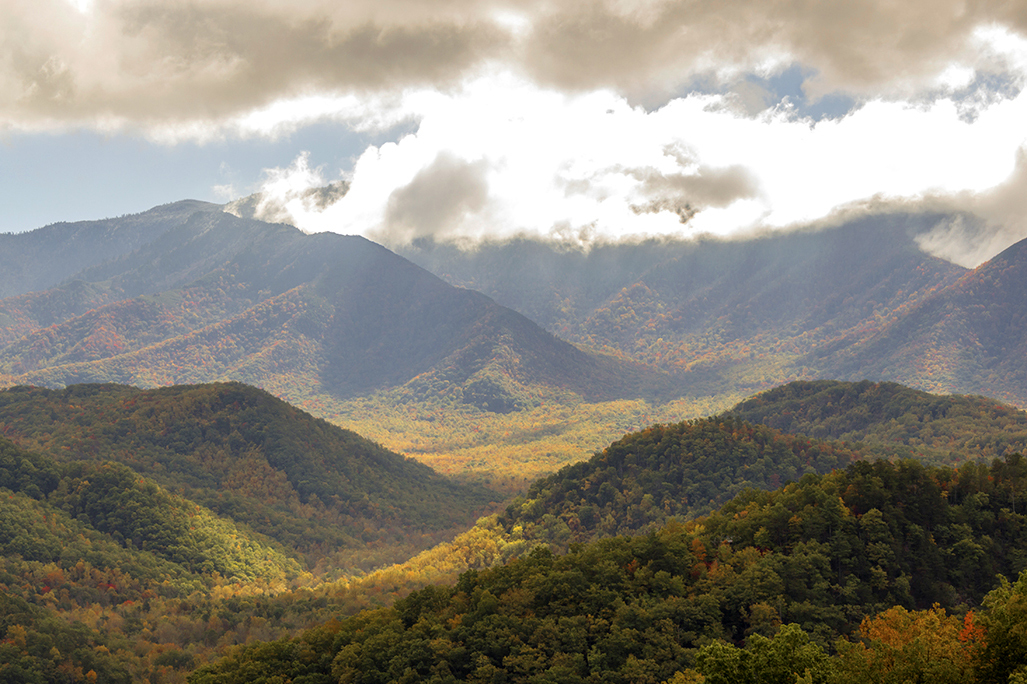 Rolling Great Smoky Mountains covered in fall foliage with sunlight breaking through clouds.